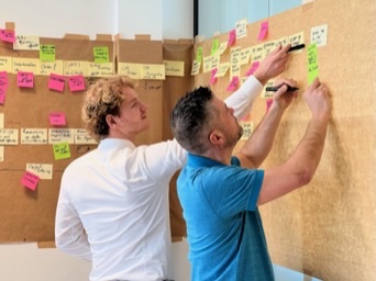 Twee mensen werken samen post-its uit op een corkboard-wand.
