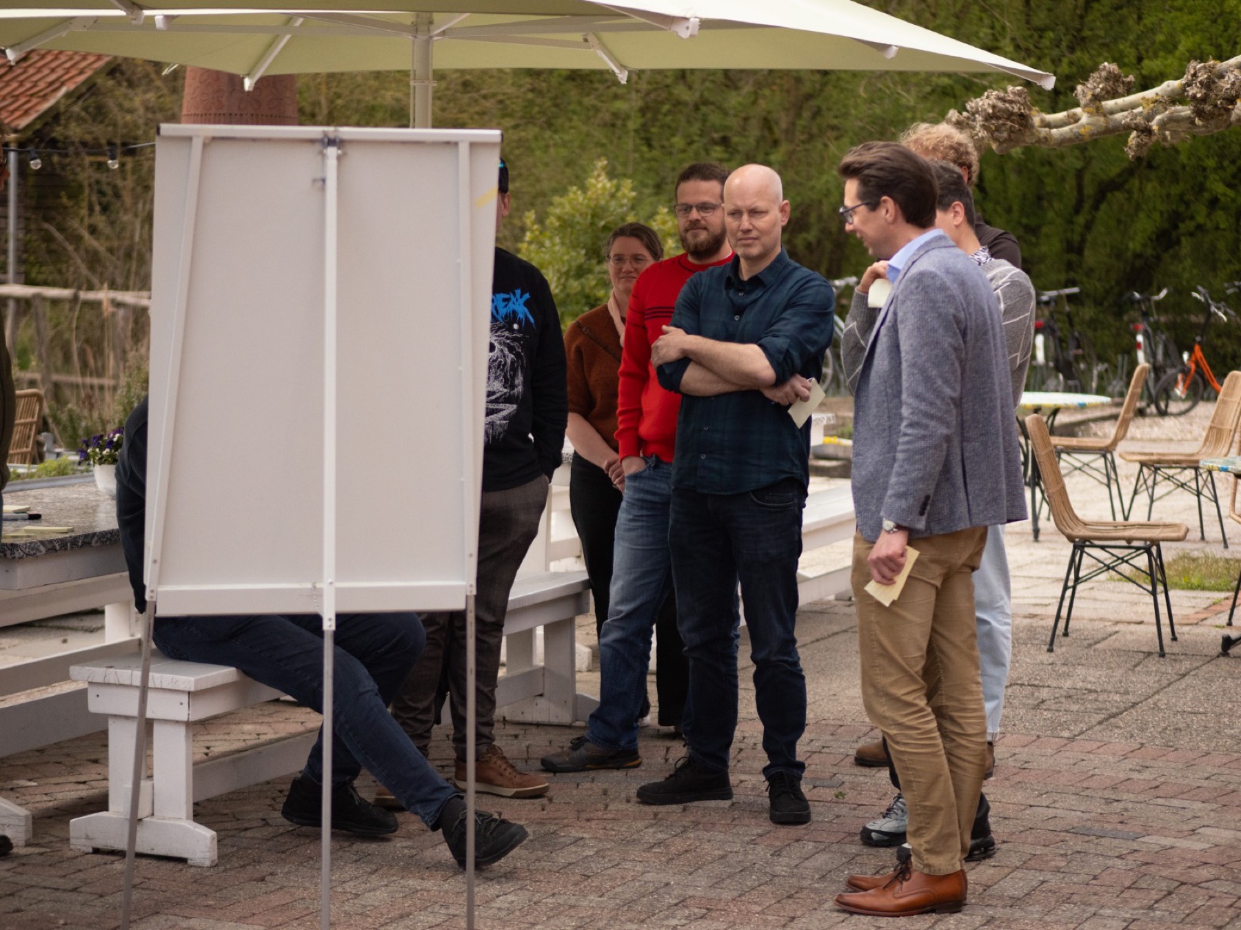 Groep collega's luistert aandachtig tijdens een workshop op een terras, met een flipchart erbij.