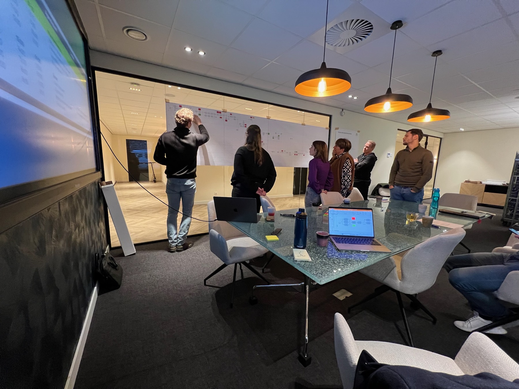 Groep collega's bespreekt een procesmuur vol post-its in een vergaderruimte.
